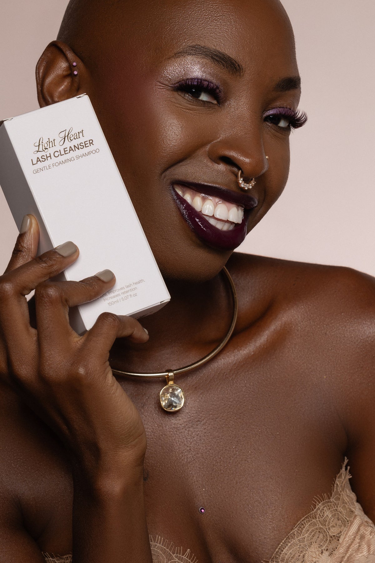 Light Heart Beauty Gentle Foaming Lash Cleanser bottle held by a smiling woman with dark skin and purple makeup.